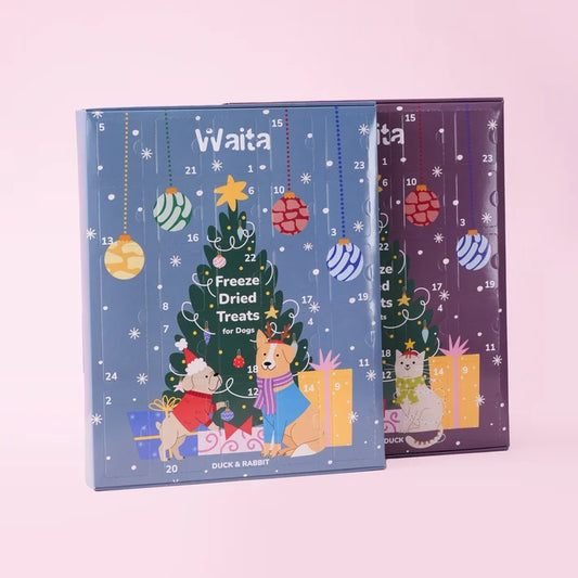 Two Christmas-themed freeze-dried pet treats advent calendars, one blue with a dog and the other purple with a cat, both decorated with a Christmas tree and ornaments, set against a solid pink background.