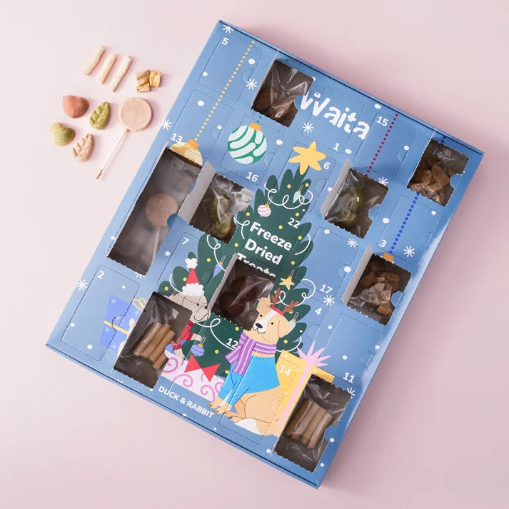 A blue Duck & Bunny Freeze Dried Advent Calendar with Christmas-themed illustrations and numbered compartments, shown on a light pink background with a few loose freeze-dried treats beside it.