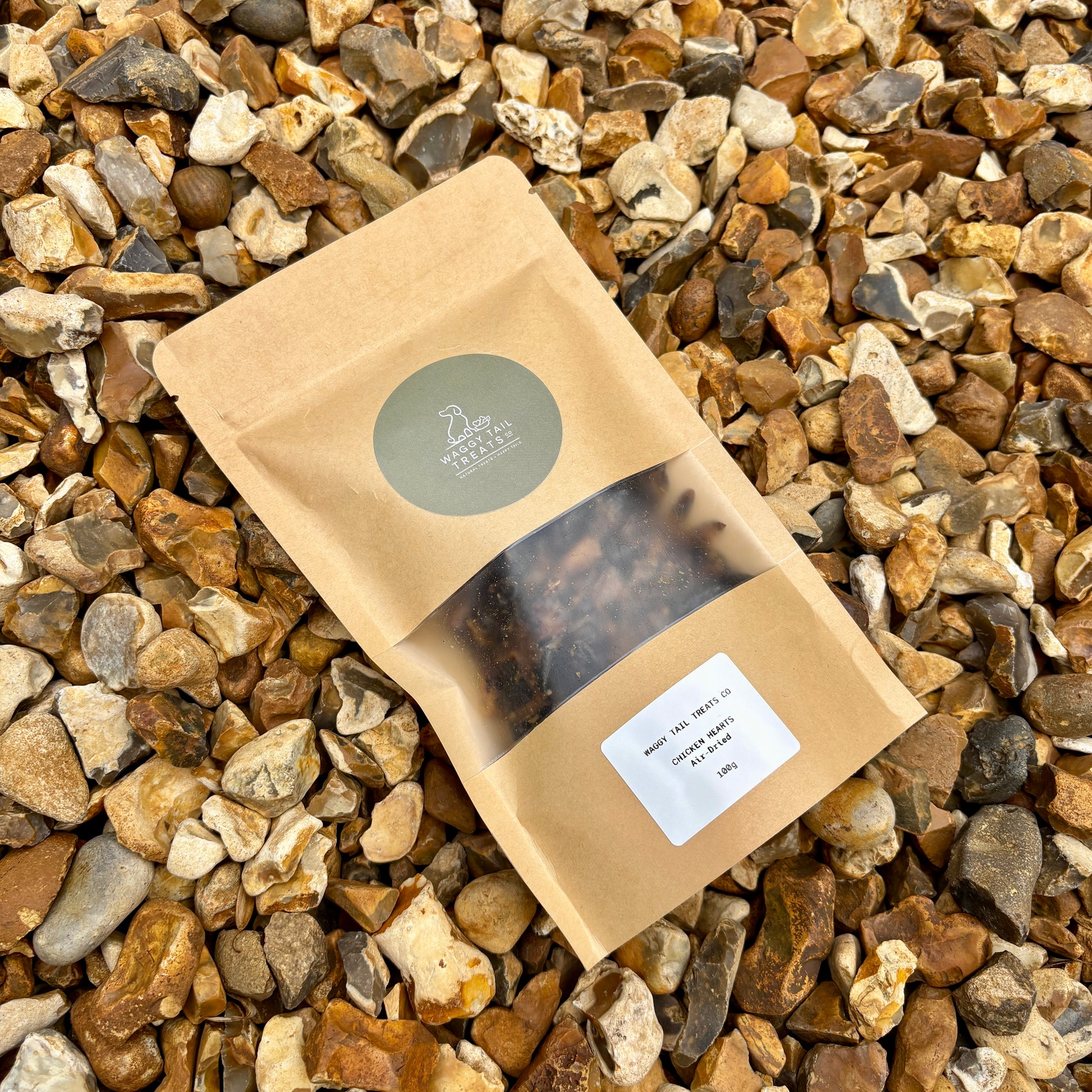 Kraft paper package with a clear window showing natural dog treats chicken hearts on a bed of small stones