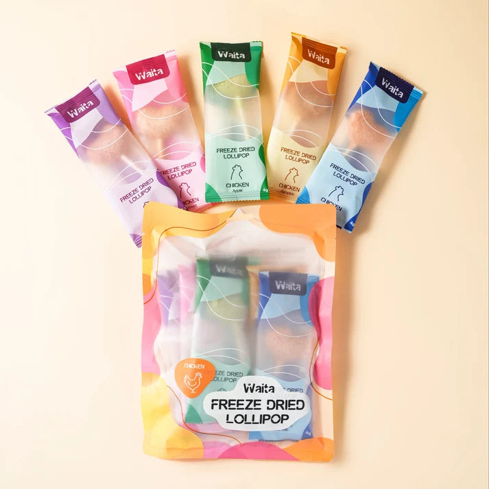 Five individually wrapped Waita freeze dried lollipop packets are arranged around a larger clear-front pouch on a warm beige background. The packets show different flavor/color options including purple, pink, green, orange, and blue, with the larger front pouch showing two lollipops inside.
